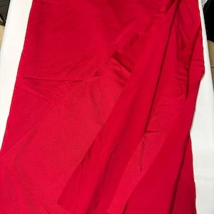 May include: A vibrant red fabric with a smooth, slightly reflective surface texture. The material is draped in soft, flowing folds, creating subtle variations in light and shadow across the solid, saturated crimson color of the textile.