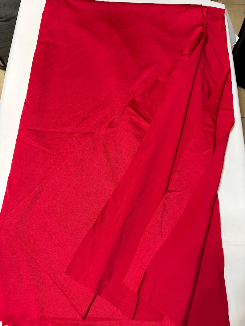 May include: A vibrant red fabric with a smooth, slightly reflective surface texture. The material is draped in soft, flowing folds, creating subtle variations in light and shadow across the solid, saturated crimson color of the textile.