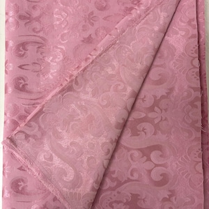 May include: A folded piece of pink fabric with a detailed, embossed floral pattern. The fabric has a soft sheen and a slightly textured surface. The edges of the fabric are frayed, adding a vintage or handmade feel. The overall aesthetic is elegant and decorative.