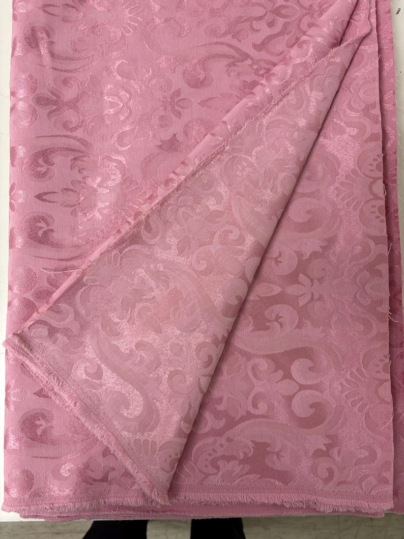 May include: A folded piece of pink fabric with a detailed, embossed floral pattern. The fabric has a soft sheen and a slightly textured surface. The edges of the fabric are frayed, adding a vintage or handmade feel. The overall aesthetic is elegant and decorative.