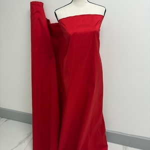 May include: A bolt of solid red fabric draped over a mannequin. The material has a smooth, slightly reflective surface and a consistent, vibrant red color throughout. The fabric is wrapped around the torso and extends down to the floor.