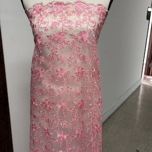 May include: Pink floral lace fabric displayed on a mannequin. The delicate lace features an intricate pattern of flowers and vines. The fabric is sheer, allowing the mannequin's form to be partially visible. The lace is a soft pink color.