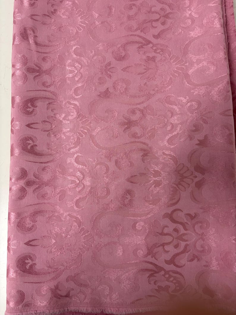 May include: A folded piece of pink fabric with a repeating floral pattern. The fabric has a subtle sheen, and the design is slightly raised, creating a textured effect. The overall look is elegant and suitable for various textile projects.