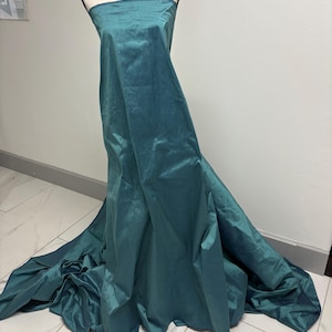 May include: A strapless floor-length gown in a deep teal color with a subtle sheen. The fabric is draped over a mannequin, creating soft folds that extend onto the floor, forming a train. The material has a smooth, slightly reflective texture.