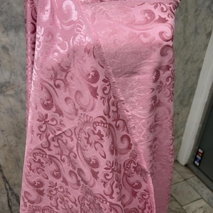 May include: A bolt of pink fabric with a detailed floral and scroll pattern. The fabric is draped over a mannequin, showcasing its texture and design. The edges of the fabric are unfinished, revealing the weave.