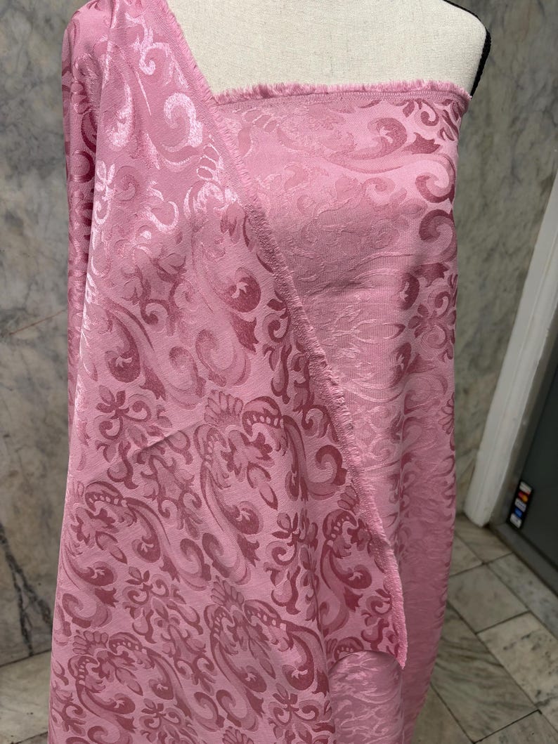 May include: A bolt of pink fabric with a detailed floral and scroll pattern. The fabric is draped over a mannequin, showcasing its texture and design. The edges of the fabric are unfinished, revealing the weave.