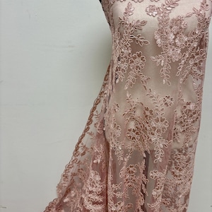 May include: A dusty rose lace fabric draped over a mannequin, featuring intricate floral embroidery and delicate sequin embellishments throughout the mesh material, with a scalloped edge detail along the top and side of the textile.