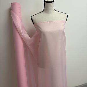 Puede incluir: Un rollo de tela rosa claro y tela drapeada sobre un maniquí. La tela es un material transparente y ligero, probablemente gasa u organza. El maniquí es un maniquí con un contorno negro.