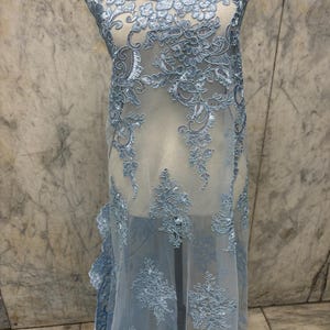 May include: Light blue lace fabric with intricate floral and scrollwork embroidery. The delicate, sheer material is draped over a mannequin, showcasing the detailed design. The lace features a scalloped edge and is suitable for bridal or formal wear.