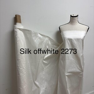 Może przedstawiać: Rolka białej jedwabnej tkaniny z napisem "Silk offwhite 2273". Tkanina jest udrapowana na manekinie. Jedwab ma subtelny połysk i wydaje się lekki. Tło to gładka biała ściana.