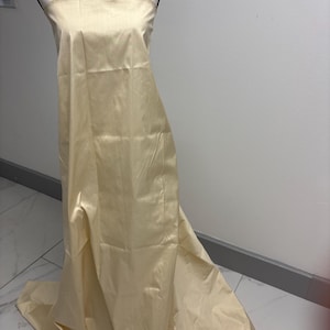 May include: A floor-length, strapless gown in a pale champagne-colored satin fabric. The garment features a smooth, lustrous finish with a straight-across neckline and a long, flowing skirt that pools slightly at the base on a mannequin form.