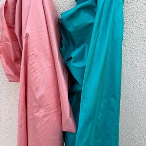 May include: Two lengths of fabric, one in a soft pink and the other in a vibrant teal, hang against a textured white wall. The fabrics are draped, showing their smooth, lustrous surfaces, ideal for sewing projects.