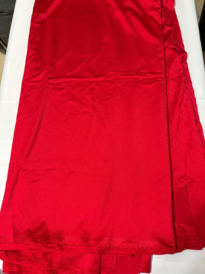 May include: A vibrant red fabric with a smooth, slightly reflective surface texture. The material is folded in layers, displaying a consistent solid crimson color throughout. The bottom edge features a subtle horizontal ribbed pattern detail.