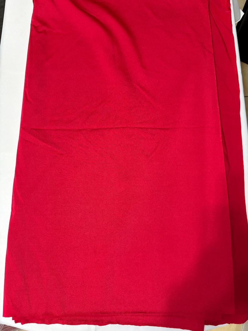 May include: A solid, vibrant red fabric with a smooth, matte texture. The material is draped in soft, gentle folds, creating subtle shadows and highlights across the surface. The color is consistent throughout, showing a deep, saturated red hue.