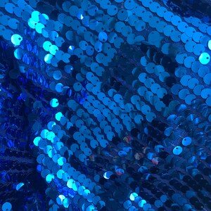 Może przedstawiać: Zbliżenie tkaniny pokrytej błyszczącymi, okrągłymi, niebieskimi cekinami. Cekiny są gęsto upakowane i ułożone w ukośnych rzędach, tworząc efekt połysku. Tło to ciemnoniebieska siatka.