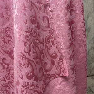 May include: Close-up of a pink fabric with a detailed damask pattern. The fabric has a textured surface and a slight sheen, with the pattern slightly darker than the base color. The edges of the fabric are visible.