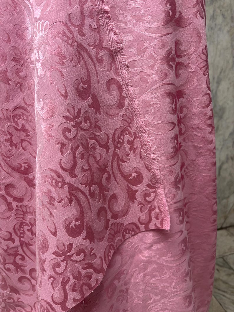 May include: Close-up of a pink fabric with a detailed damask pattern. The fabric has a textured surface and a slight sheen, with the pattern slightly darker than the base color. The edges of the fabric are visible.
