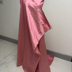 May include: A length of solid pink fabric draped over a dress form. The material has a smooth, lustrous finish with a subtle sheen, creating soft folds and cascading layers that extend toward the floor, showcasing the texture and color.