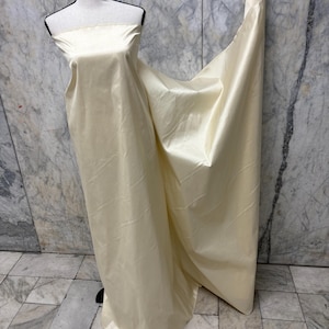 May include: A cream-colored fabric draped over a mannequin. The fabric appears to be a smooth, flowing material, possibly silk or satin, with a subtle sheen. The fabric is unhemmed and appears to be a continuous length, suitable for dressmaking or other textile projects.