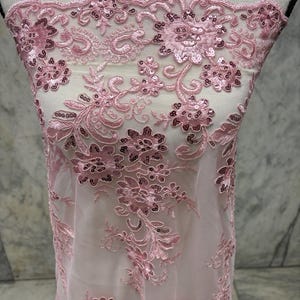 May include: Pale pink floral lace fabric with sequin embellishments. The sheer fabric displays embroidered flowers and ornate patterns. Ideal for dresses, veils, or other clothing items, the lace has a delicate, intricate design.