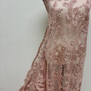 Può includere: Tessuto in pizzo rosa antico con ricami floreali. Il delicato pizzo presenta un bordo smerlato e motivi intricati di fiori e foglie. Il materiale trasparente è drappeggiato su un manichino, mostrando il flusso e la consistenza del tessuto.