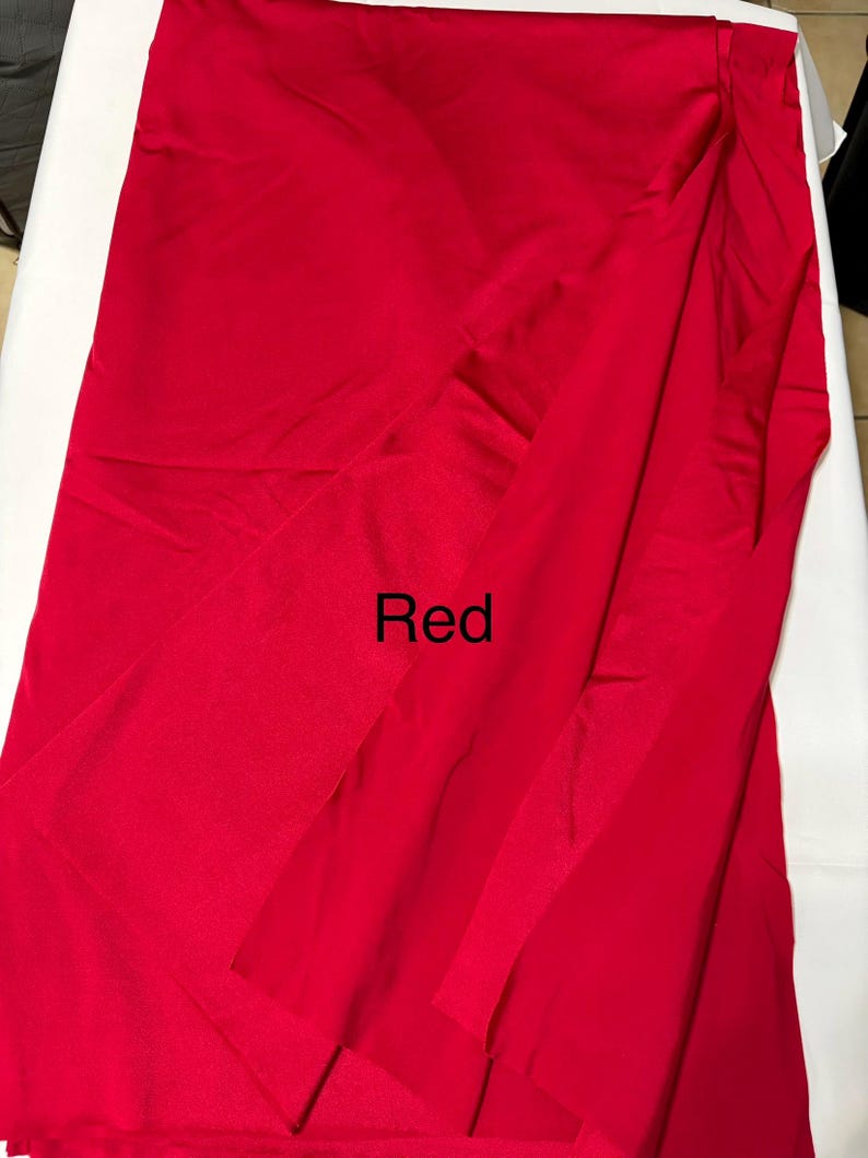 May include: A solid, vibrant red fabric with a smooth, slightly reflective surface texture. The material is draped in soft, vertical folds, creating natural shadows and highlights across the surface. The word Red is printed in black text.