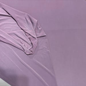 May include: A solid mauve-colored fabric with a smooth, matte finish. The material is draped to show its soft, flexible texture, with gentle folds creating subtle shadows across the surface, highlighting the consistent, muted purple tone throughout.
