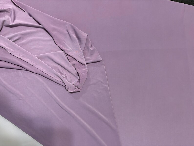 May include: A solid mauve-colored fabric with a smooth, matte finish. The material is draped to show its soft, flexible texture, with gentle folds creating subtle shadows across the surface, highlighting the consistent, muted purple tone throughout.