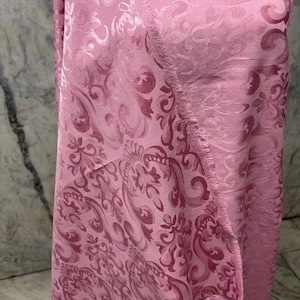 May include: A pink fabric with a detailed, swirling floral pattern. The fabric has a satin sheen and a soft texture. The design features a repeating motif of stylized flowers and leaves in varying shades of pink. The edges of the fabric are visible, showing a fringed detail.