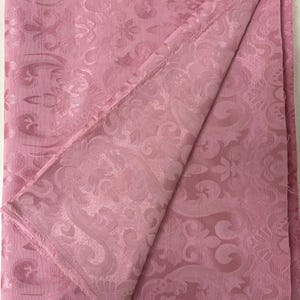 May include: A folded piece of pink fabric with a detailed, embossed floral pattern. The fabric has a soft, matte finish and a visible woven texture. The edges of the fabric are unfinished, revealing the raw weave. The overall aesthetic is elegant and decorative.