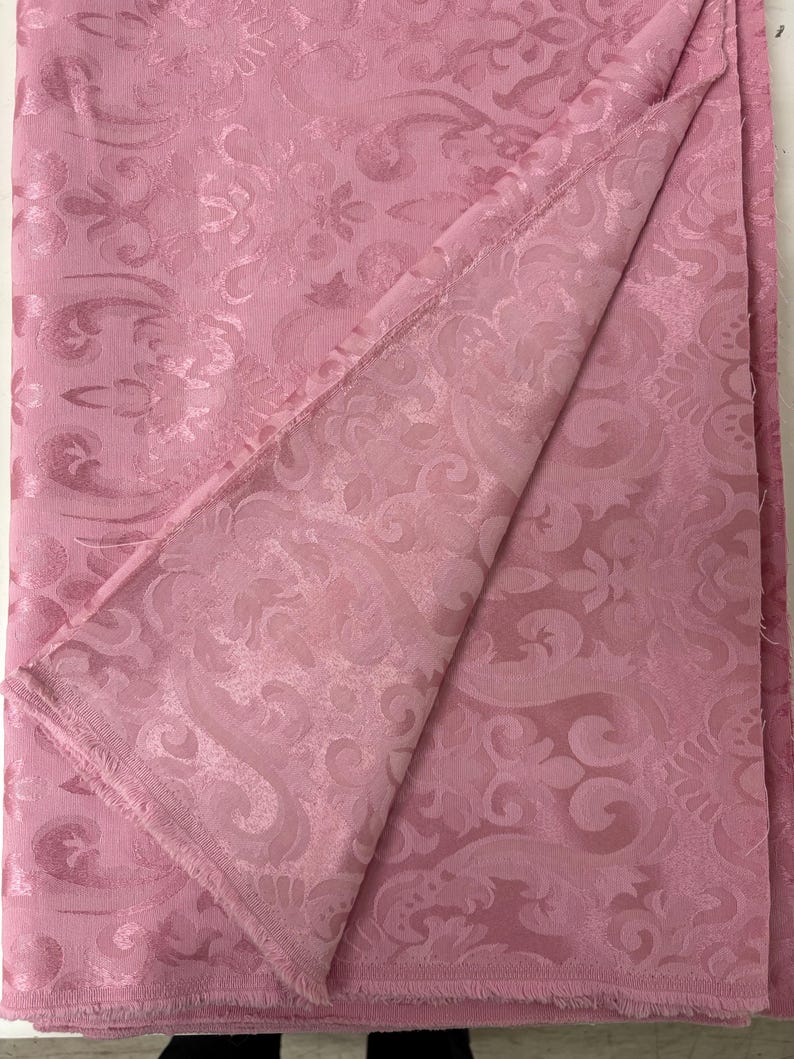 May include: A folded piece of pink fabric with a detailed, embossed floral pattern. The fabric has a soft, matte finish and a visible woven texture. The edges of the fabric are unfinished, revealing the raw weave. The overall aesthetic is elegant and decorative.