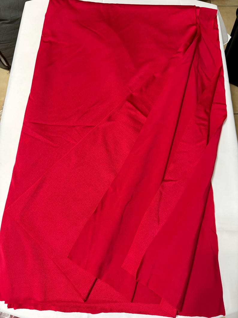 May include: A vibrant red fabric with a smooth, slightly reflective surface. The material is draped in a way that creates soft folds and layers, showcasing the consistent, solid red color throughout the entire piece of textile.