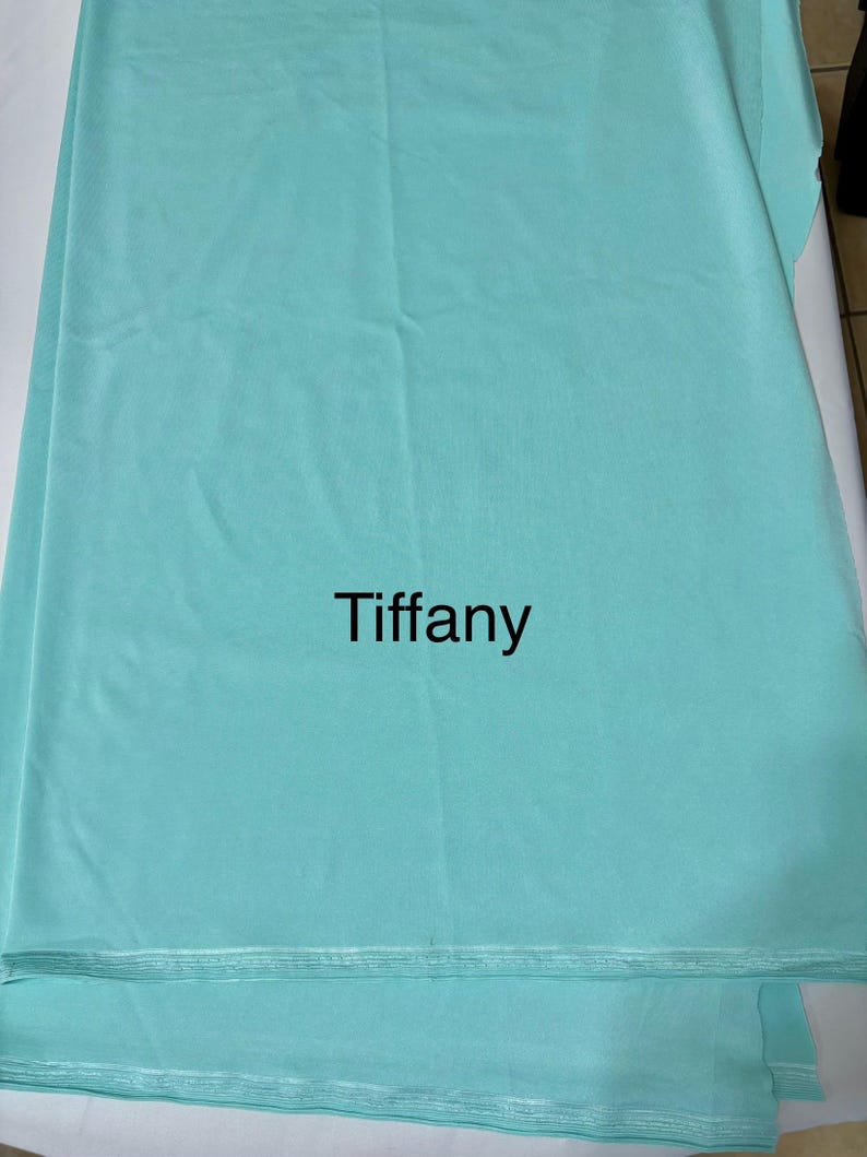 May include: A solid light turquoise fabric featuring the word Tiffany printed in black in the center. The material has a smooth, matte texture with visible horizontal folds and a finished edge at the bottom, displaying a consistent color throughout.