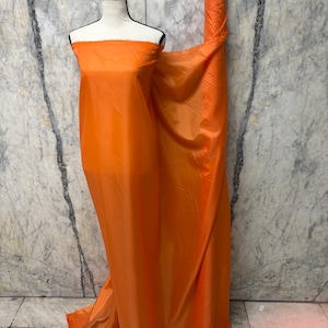 May include: Bright orange fabric draped over a mannequin and rolled up. The fabric has a shiny, smooth texture and is a vibrant, solid color. The background is a marble-like wall.