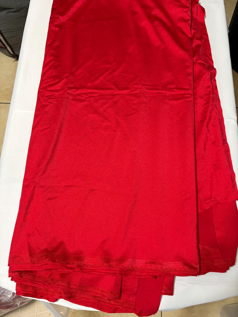 May include: A solid, vibrant red fabric with a smooth, slightly reflective surface texture. The material is folded in layers, showing a consistent, deep crimson color throughout the entire piece of textile, which appears to be a single, continuous length.