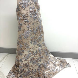 May include: A beige fabric with a floral pattern in silver and blue. The fabric is draped over a mannequin, showcasing the detailed design and texture. The floral pattern includes leaves and branches.