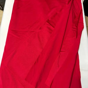 May include: A solid, vibrant red fabric with a smooth, slightly reflective finish. The material is draped in soft, vertical folds, creating a layered appearance that highlights the consistent, deep crimson color across the entire surface.