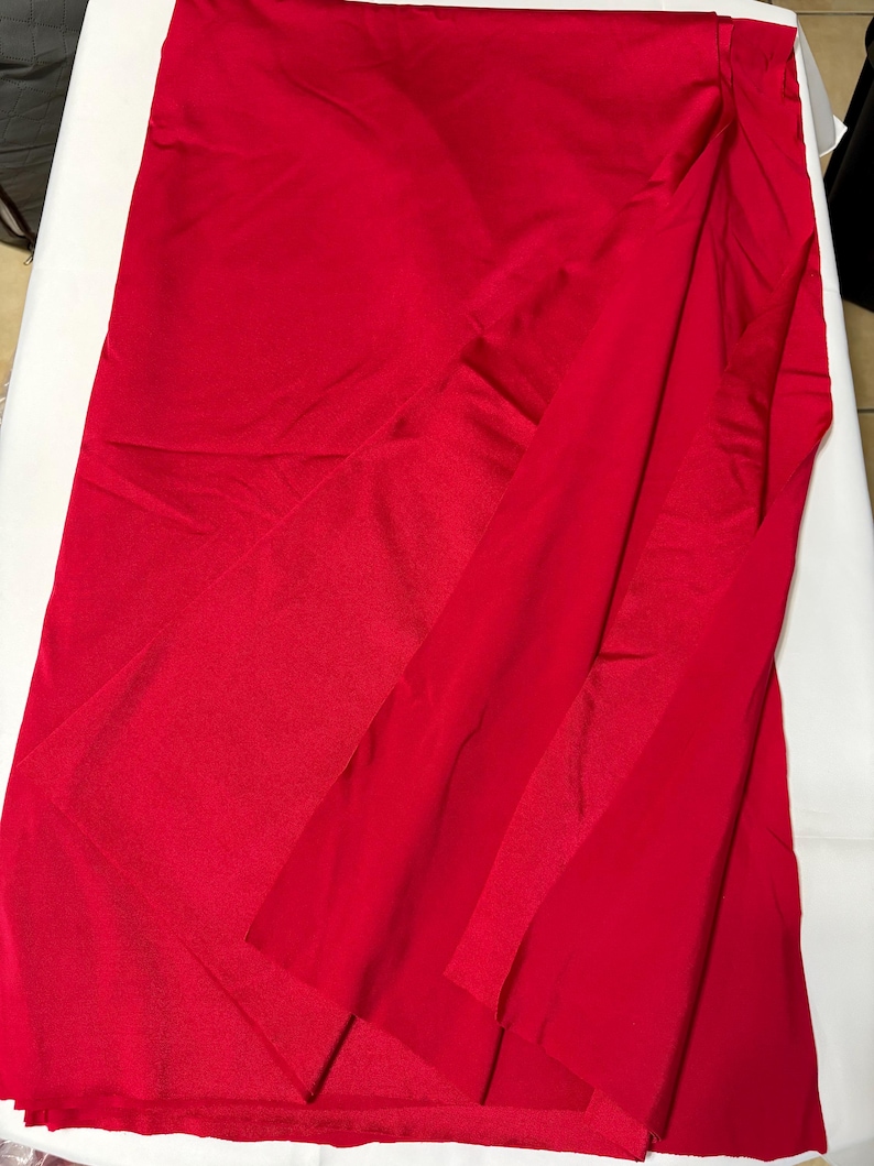 May include: A solid, vibrant red fabric with a smooth, slightly reflective finish. The material is draped in soft, vertical folds, creating a layered appearance that highlights the consistent, deep crimson color across the entire surface.