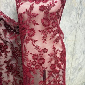 Op de afbeelding: Bordeaux bloemen kanten stof. De transparante stof heeft een ingewikkeld geborduurd ontwerp van bloemen en bladeren. De geschulpte rand voegt een decoratief tintje toe. De stof wordt op een mannequin getoond, waarbij de drapering en textuur worden getoond.