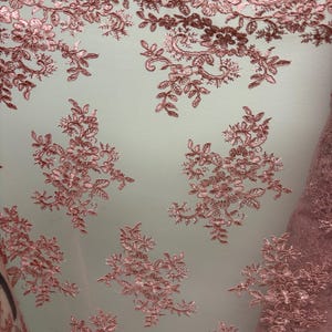 K&ouml;nnte beinhalten: Nahaufnahme eines zarten, rosafarbenen Spitzenstoffs. Die Spitze weist komplizierte Blumen- und Blattmuster mit einem gewellten Rand auf. Der Stoff ist halbtransparent und zeigt einen hellen Hintergrund. Ideal f&uuml;r Brautmode oder Dekorationsprojekte.