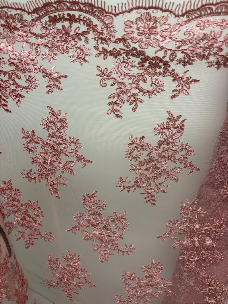 K&ouml;nnte beinhalten: Nahaufnahme eines zarten, rosafarbenen Spitzenstoffs. Die Spitze weist komplizierte Blumen- und Blattmuster mit einem gewellten Rand auf. Der Stoff ist halbtransparent und zeigt einen hellen Hintergrund. Ideal f&uuml;r Brautmode oder Dekorationsprojekte.