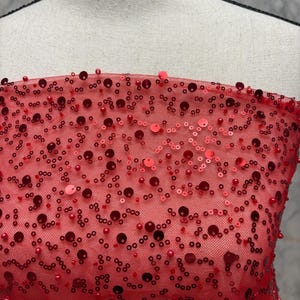 May include: Close-up of red fabric decorated with sequins and beads. The fabric is a sheer mesh, with various sizes of red sequins and beads scattered across the surface. The sequins and beads create a textured, shimmering effect. The fabric is draped over a mannequin.
