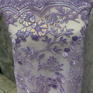 May include: Close-up of lavender lace fabric with floral and leaf patterns. The lace features embroidered flowers with sequin centers and a scalloped edge. The fabric is semi-sheer, revealing a light background.