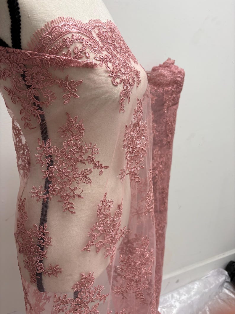 K&ouml;nnte beinhalten: Staubrosa Spitzenstoff mit floraler Stickerei. Der zarte Stoff ist &uuml;ber eine Schaufensterpuppe drapiert und zeigt die komplizierten Details des Designs. Die Spitze hat einen gewellten Rand und einen transparenten Hintergrund.