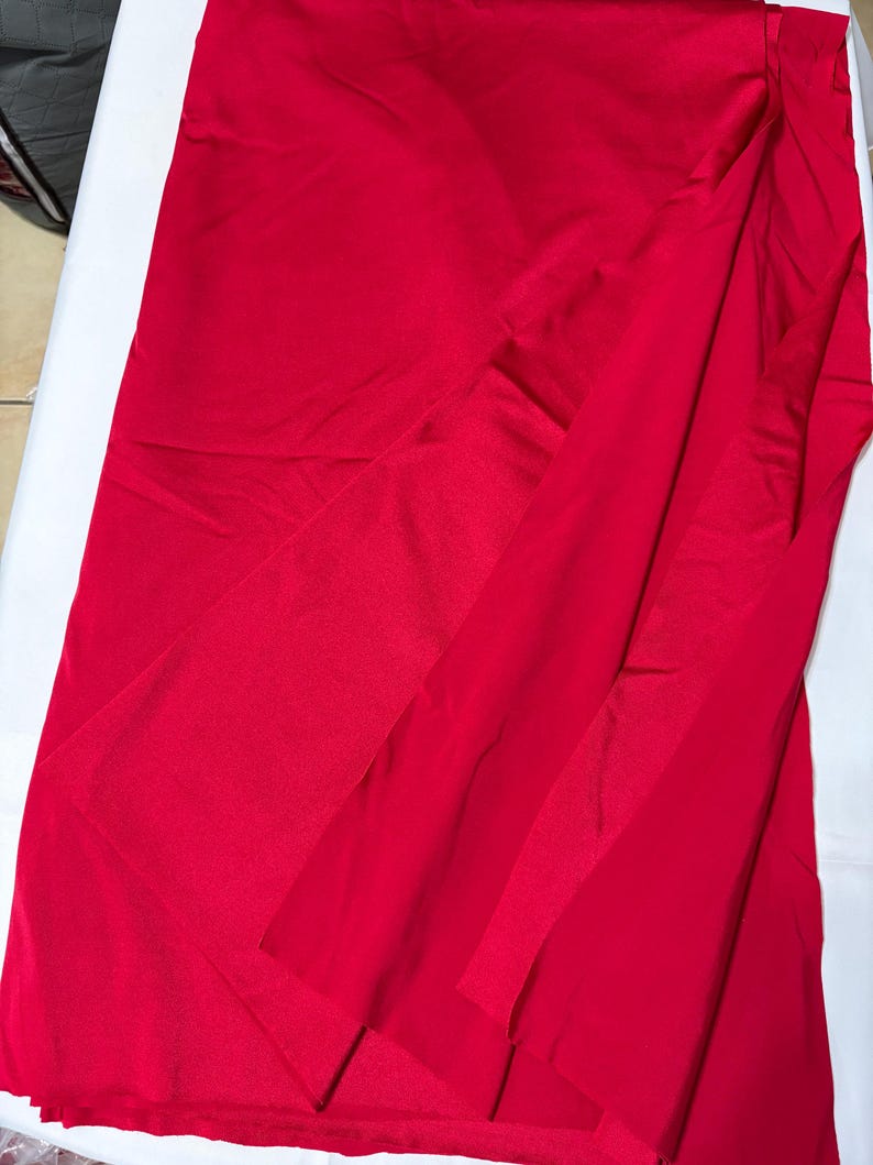 May include: A vibrant red fabric with a smooth, slightly reflective surface texture. The material is draped in a way that creates soft, flowing folds and layers, showcasing the consistent, solid red color across the entire visible surface area.