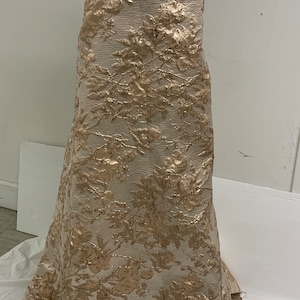 May include: A white fabric with a gold floral pattern. The fabric is draped over a dress form.