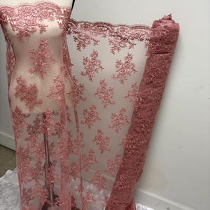 K&ouml;nnte beinhalten: Staubrosa Spitzenstoff mit floraler Stickerei. Der Stoff ist &uuml;ber eine Schaufensterpuppe drapiert und aufgerollt, wodurch die komplizierten Details und die transparente Qualit&auml;t zur Geltung kommen. Die Spitze hat einen gewellten Rand und ein zartes Muster.