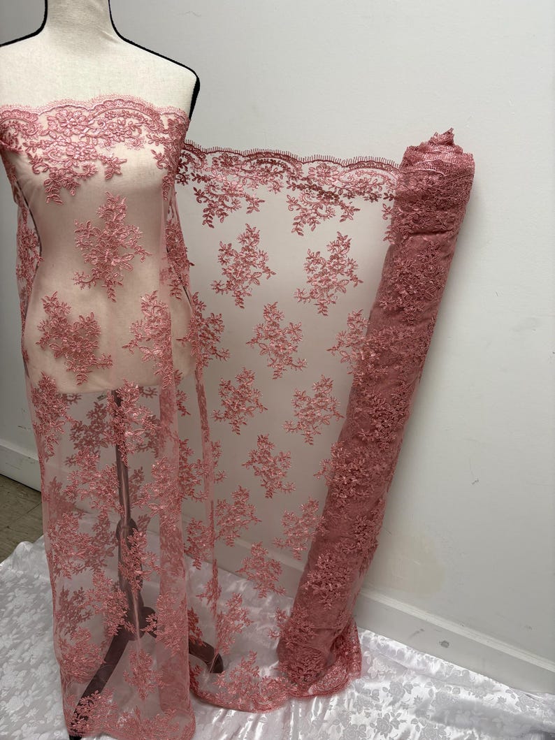 K&ouml;nnte beinhalten: Staubrosa Spitzenstoff mit floraler Stickerei. Der Stoff ist &uuml;ber eine Schaufensterpuppe drapiert und aufgerollt, wodurch die komplizierten Details und die transparente Qualit&auml;t zur Geltung kommen. Die Spitze hat einen gewellten Rand und ein zartes Muster.