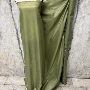 May include: Olive green fabric draped over a mannequin and rolled up. The fabric has a slight sheen and appears to be a lightweight material. The background is a marble-like wall. The fabric is suitable for apparel or home decor.