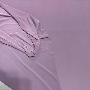 May include: A solid, light purple fabric with a smooth, matte texture. The material is draped on one side, showing soft folds and a consistent, opaque color throughout the surface. The fabric appears lightweight and flexible with a clean finish.